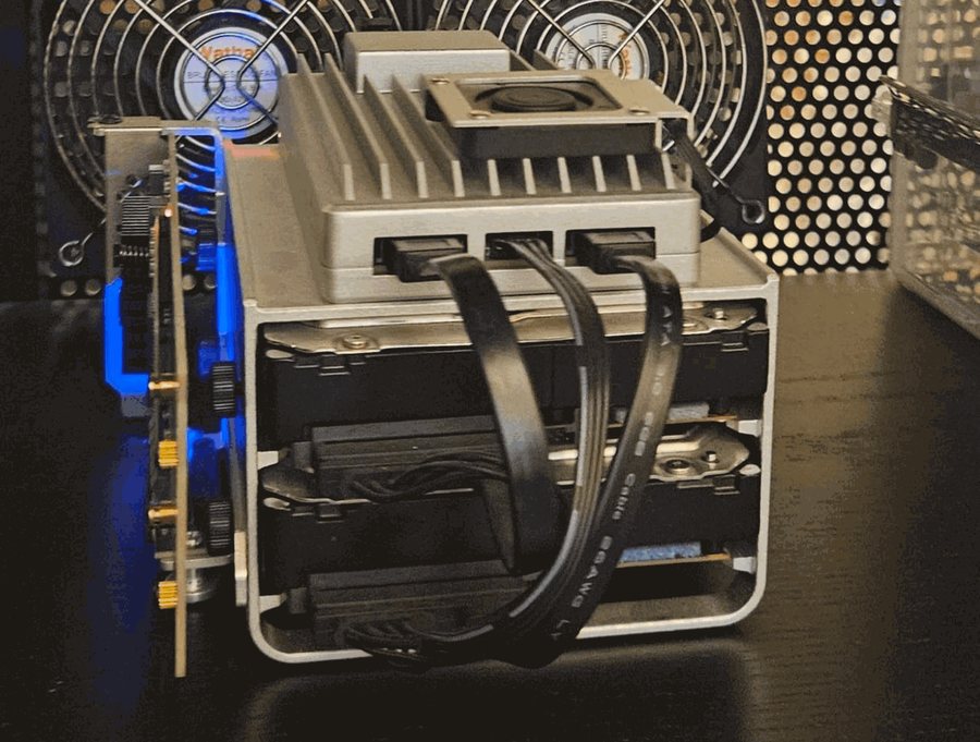 The ZimaBoard sitting on its drive cage, which holds two hard drives, with a PCIe adapter card standing on its side against the cage, lit blue